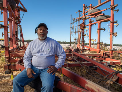 A Black farmer leans against a tractor at a cotton farm.