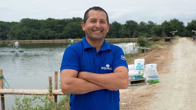 A man stands with his arms crossed wearing a blue collared shirt in front of a lake. 