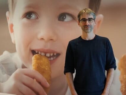 A man stands in front of a large picture of a boy eating a chicken nugget. 