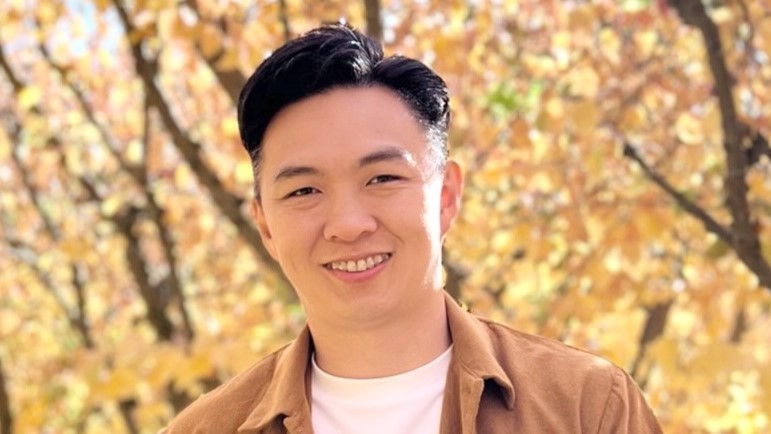 A man smiles while standing in front of trees with yellow leaves. 