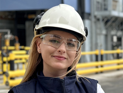 A woman wearing a hard hat and safety glasses stands outside an operations facility. A woman wearing a hard hat and safety glasses stands outside an operations facility.