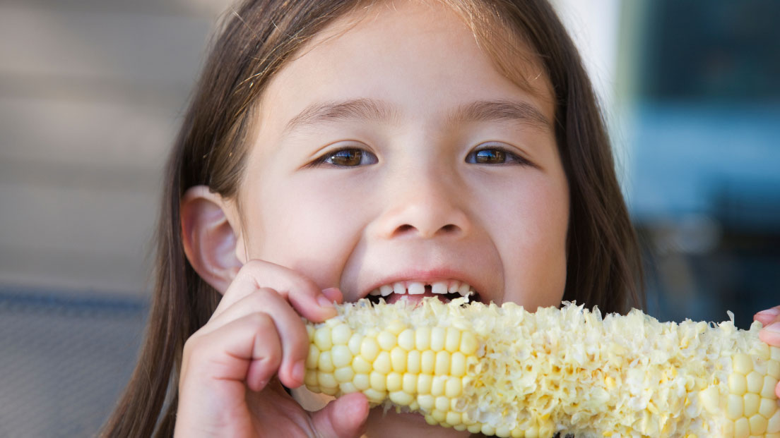 A young girl takes a bite of an ear of corn.