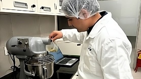 A man in a white lab coat pours Brown liquid into a stand-up mixer.