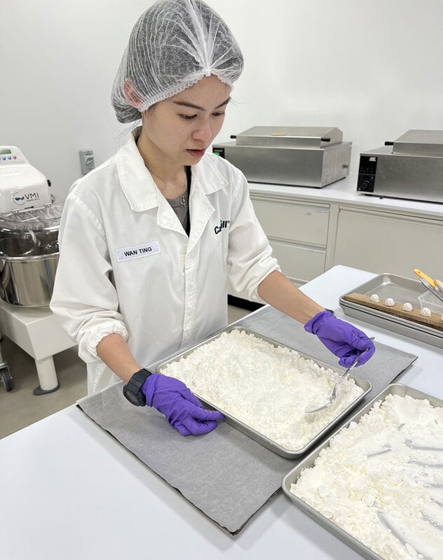 A woman in a white lab coat looks down while standing in a food science lab.