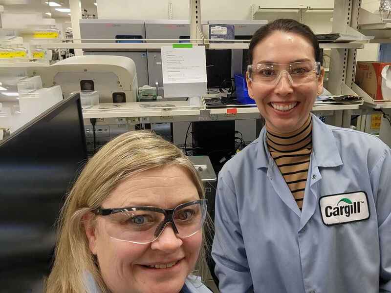 Two women in blue Cargill lab coats smile as they stand in front of a microbiome sequencer.