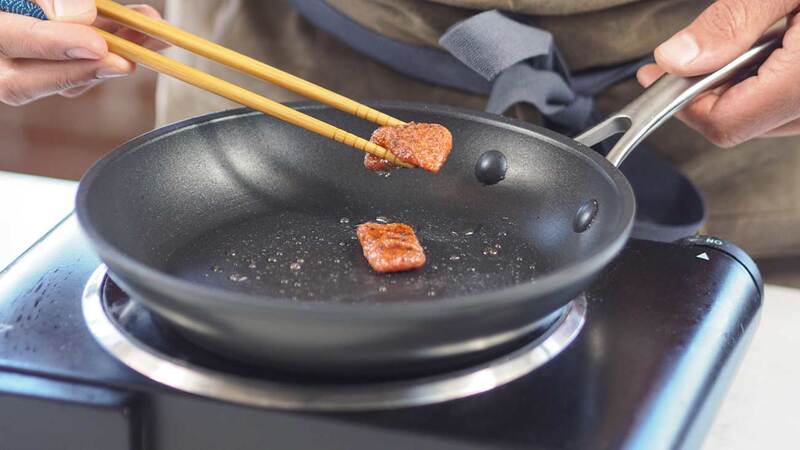 A person holds a piece of cultivated salmon between two chopsticks over a frying pan.