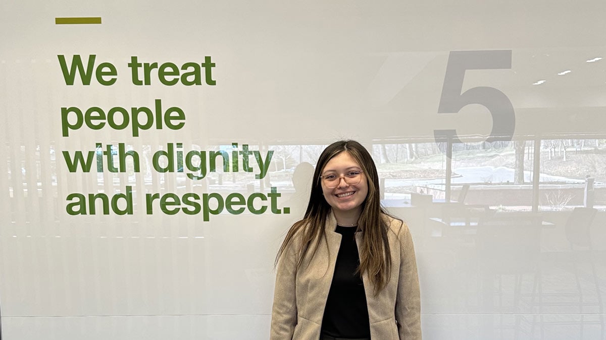 A young woman smiles in front of a wall with the message that says, ‘We treat people with dignity and respect.’