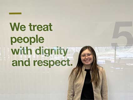 A young woman smiles in front of a wall with the message that says, ‘We treat people with dignity and respect.’ A young woman smiles in front of a wall with the message that says, ‘We treat people with dignity and respect.’