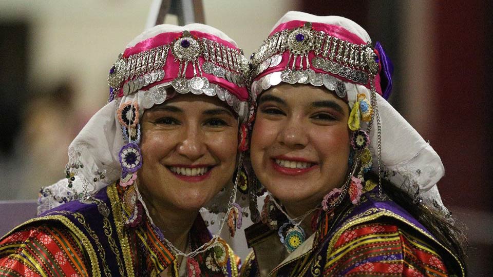 Two women smile at the camera while donning traditional Turkish garments. 