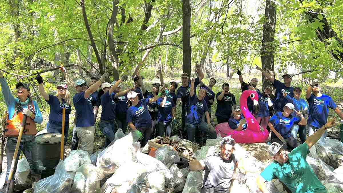 A large group of people surrounded by trees smile next to dozens of bags of recently removed trash. A large group of people surrounded by trees smile next to dozens of bags of recently removed trash.
