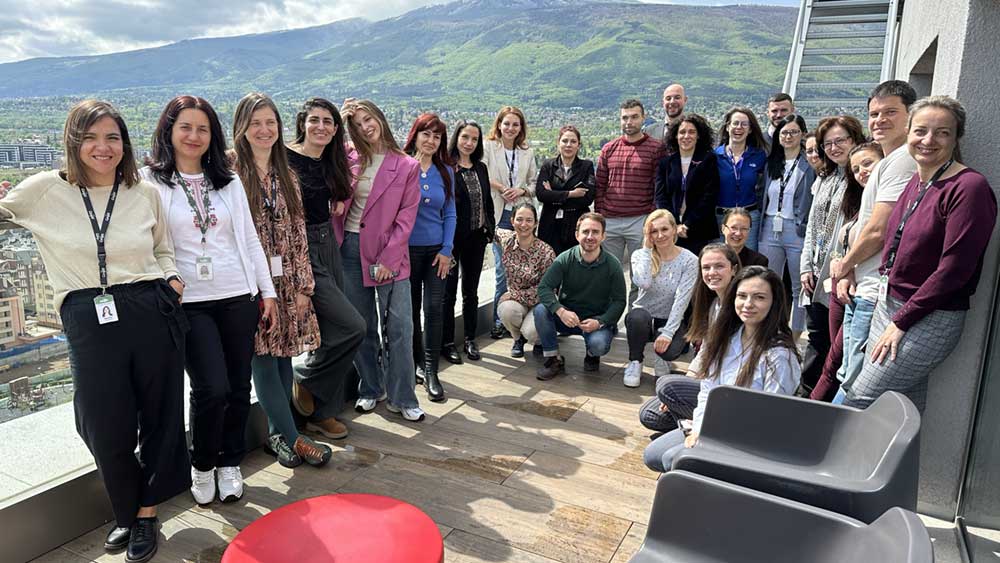 A group of employees pose for the camera at an office setting, with mountains in the background. A group of employees pose for the camera at an office setting, with mountains in the background.