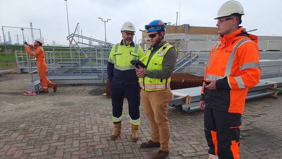 Four men in white hard hats and yellow and orange vests pilot a drone. Four men in white hard hats and yellow and orange vests pilot a drone.