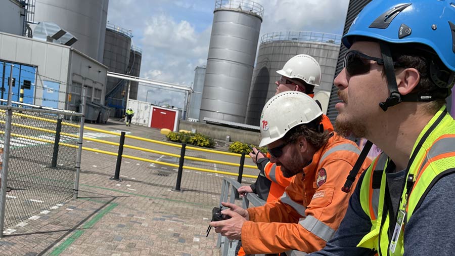 Three men in hard hats lean down and look up to the sky to see a drone flying. Three men in hard hats lean down and look up to the sky to see a drone flying.