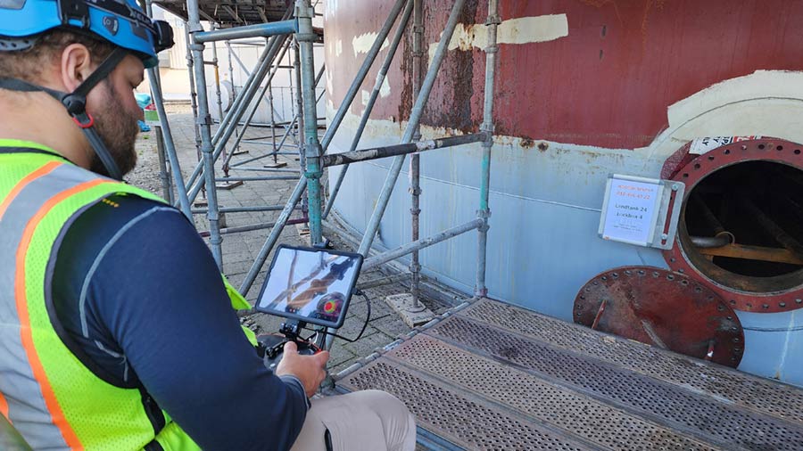 A man in a blue helmet looks down at a tablet with the view of a camera mounted on a flying drone. A man in a blue helmet looks down at a tablet with the view of a camera mounted on a flying drone.
