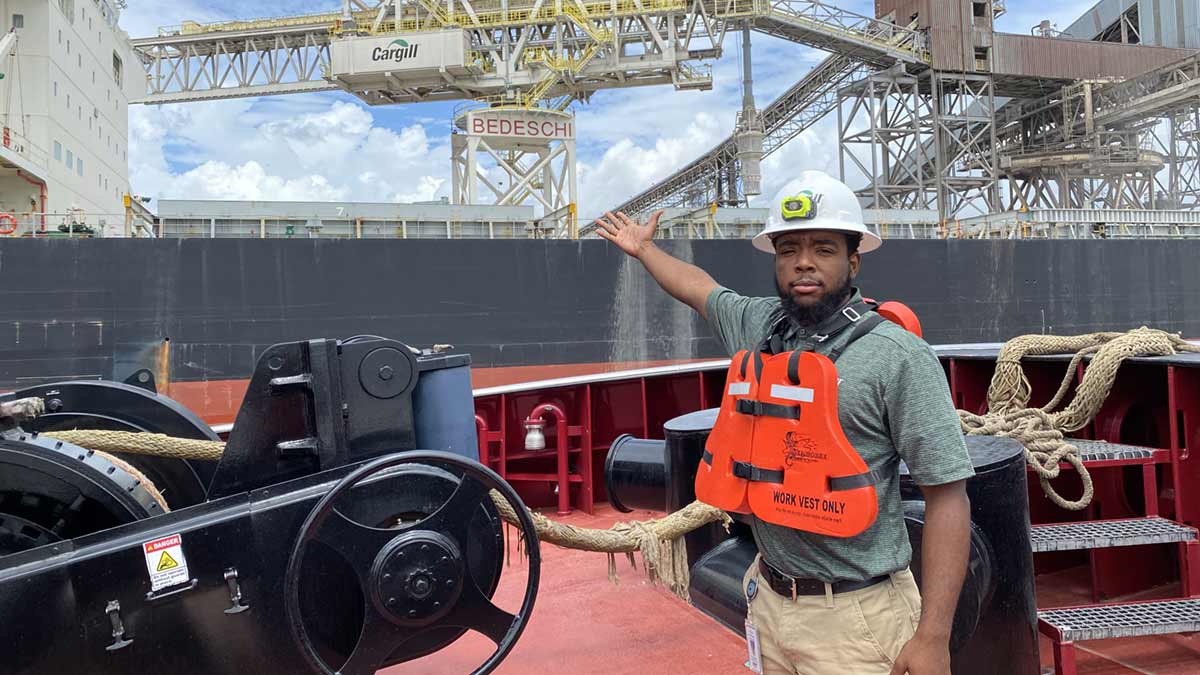 Ernest Dixon III, wearing a hard hat and safety vest, standing on a ship and pointing towards a Cargill grain facility.