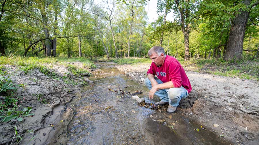 A farmer crouches down near a stream on his farmland.
