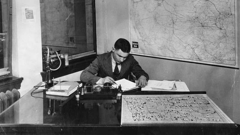 A white man in a business suit works at a large desk with papers spread in front of him. A white man in a business suit works at a large desk with papers spread in front of him.