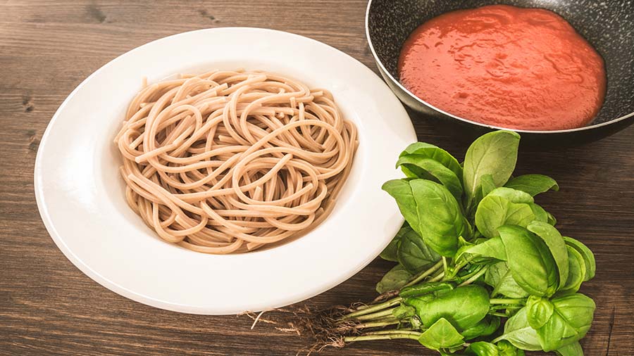 Pasta in a white bowl and red pasta sauce in a black bowl with some fresh basil on a table. 