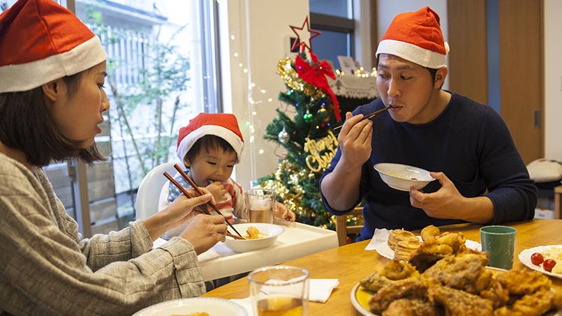 A woman, man and small child all wearing red holiday hats sit at a table with fried chicken and biscuits on it. 