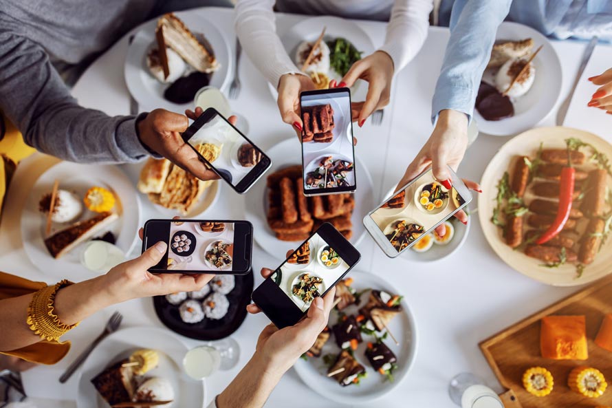 People snapping photos of food with smartphones around a table, showcasing plates filled with protein options like chicken and beef.
