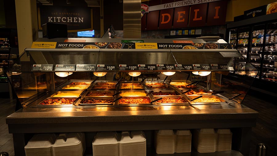 A grocery store hot food bar with trays of various cooked dishes under heat lamps, labeled with prices, and stacks of takeout containers below.
