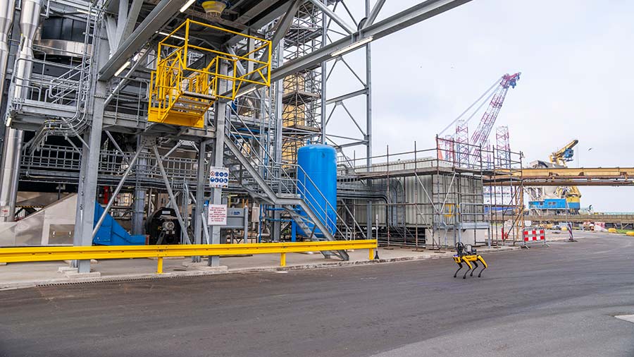 Spot the robotic dog walks on pavement outside a large operations facility.