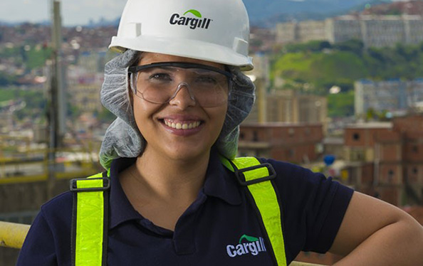 An employee in a white hard hat and yellow safety jacket stands over Spot the robotic dog as it sits on the ground in a plant. 