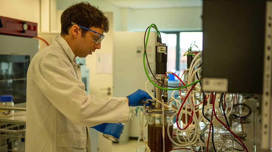 A man in a white lab coat wearing blue gloves works with a clear container of fermented liquid. 