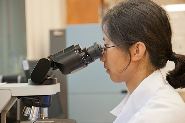 A woman wearing glasses and a white lab coat looks into a microscope. 
