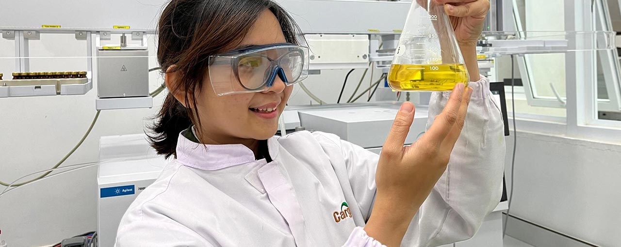 A woman wearing a white lab coat and safety glasses holds up a glass beaker with yellow liquid inside. 
