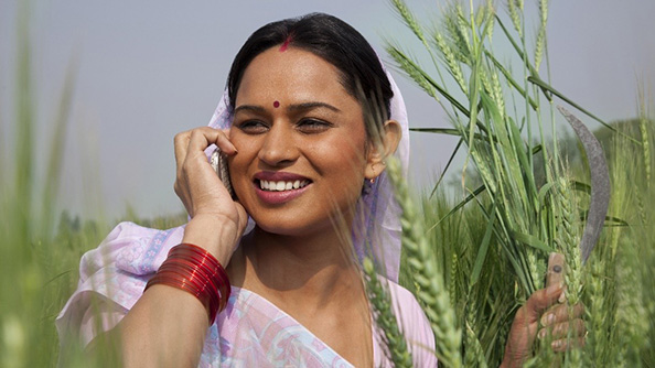 A woman standing next to a wheat field speaks into a cell phone. 