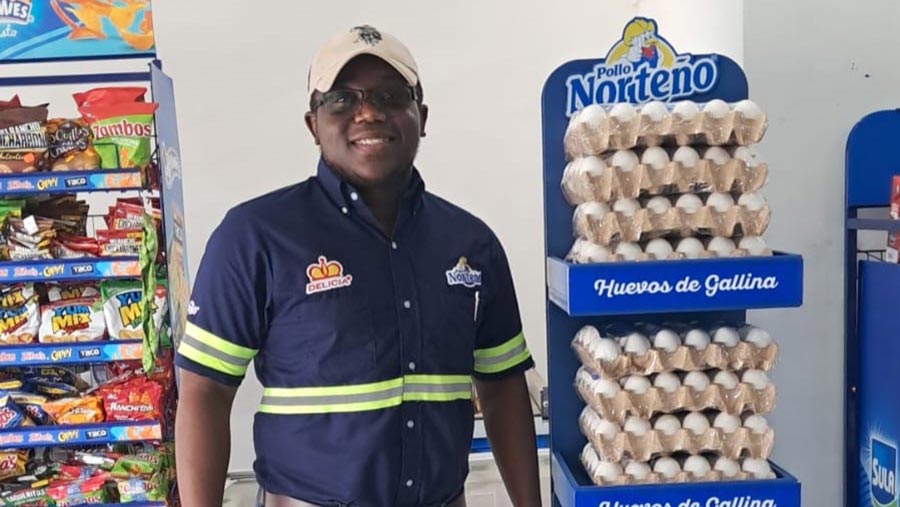 A male employee in a blue shirt and gray hat stands next to a stand in a grocery store with dozens of egg cartons. 