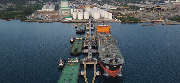 Several ships are docked in the water in front of a large refinery facility. 
