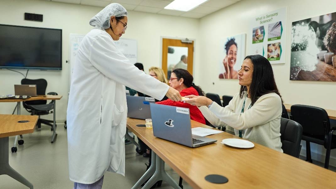 A woman in a white lab coat and white hair net collects a plate of protein chips from a woman in a brown sweater during a taste test. 