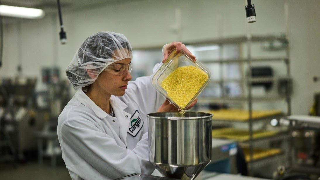 A Cargill scientist in white lab coat and hair net pours a large container of yellow protein powder into a silver machine. A Cargill scientist in white lab coat and hair net pours a large container of yellow protein powder into a silver machine.