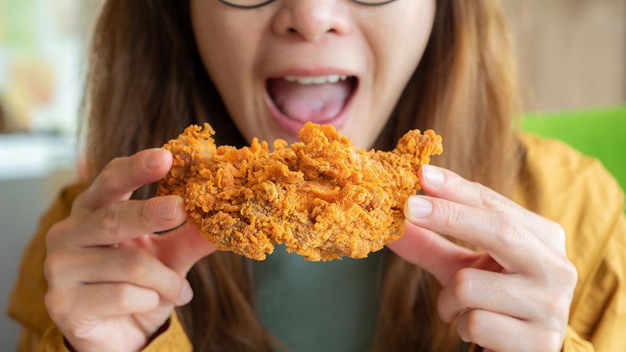 A woman holds a large piece of fried chicken in front of her mouth before taking a bite. 