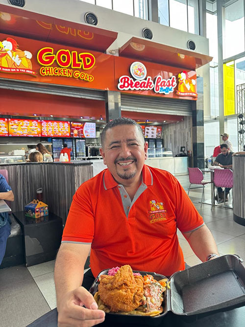 A person in a white suit and green headwrap and face mask holds a piece of chicken and takes noes while standing in a lab.