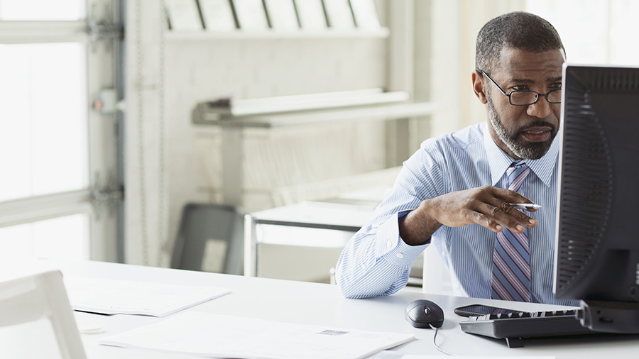 A man in a blue shirt, tie and glasses looks closely at a computer screen.  