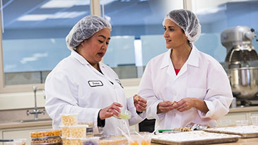 Two Cargill sensory scientists work in a lab with several products laid out in front of them on a test kitchen table. 