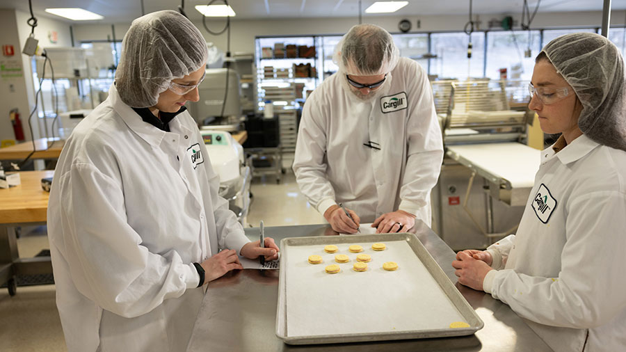 Three individuals in white coats collaborate in a professional kitchen setting, preparing food and managing tasks.