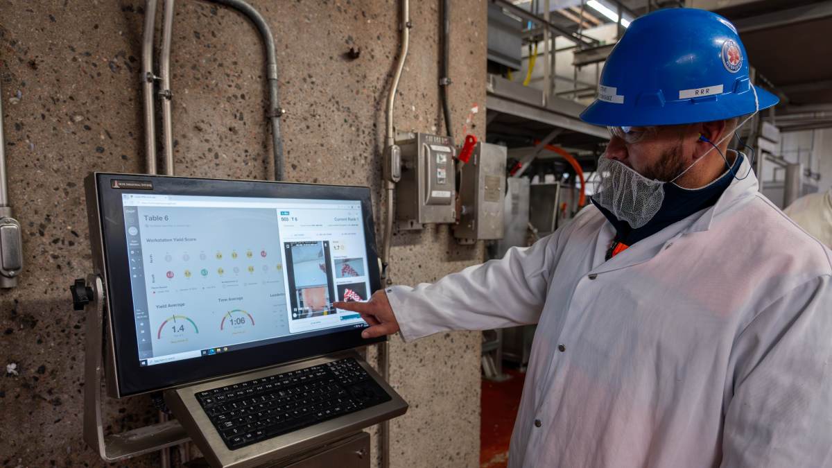 Worker in protective gear using a touchscreen monitor to analyze production data in a food processing facility.
