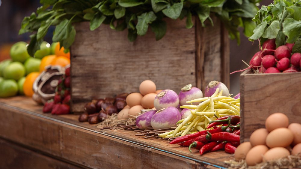 Fresh vegetables, fruits and eggs displayed on a rustic market table, including leafy greens, radishes, turnips, beans and peppers.