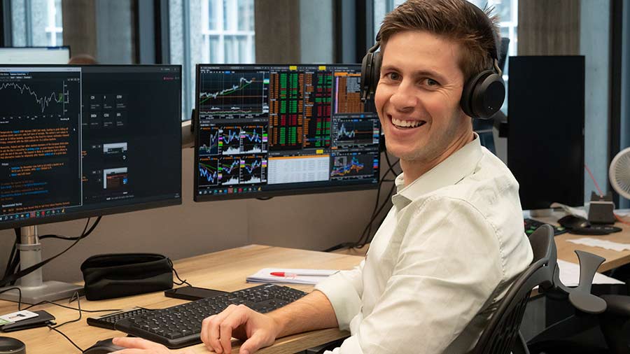 A male Cargill trader smiles at the camera while sitting in front of two computer screens with graphs and trading information on them. A male Cargill trader smiles at the camera while sitting in front of two computer screens with graphs and trading information on them.