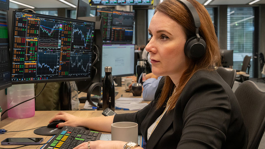 A female trader wearing black headphones and a black suit top focuses on two computer screens with financial figures and graphs. A female trader wearing black headphones and a black suit top focuses on two computer screens with financial figures and graphs.