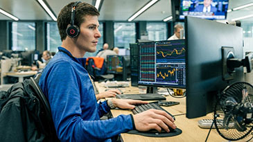 A male trader wearing black headphones scrolls with his computer mouse while looking at three computer screens on his desk inside the Cargill trading room. 