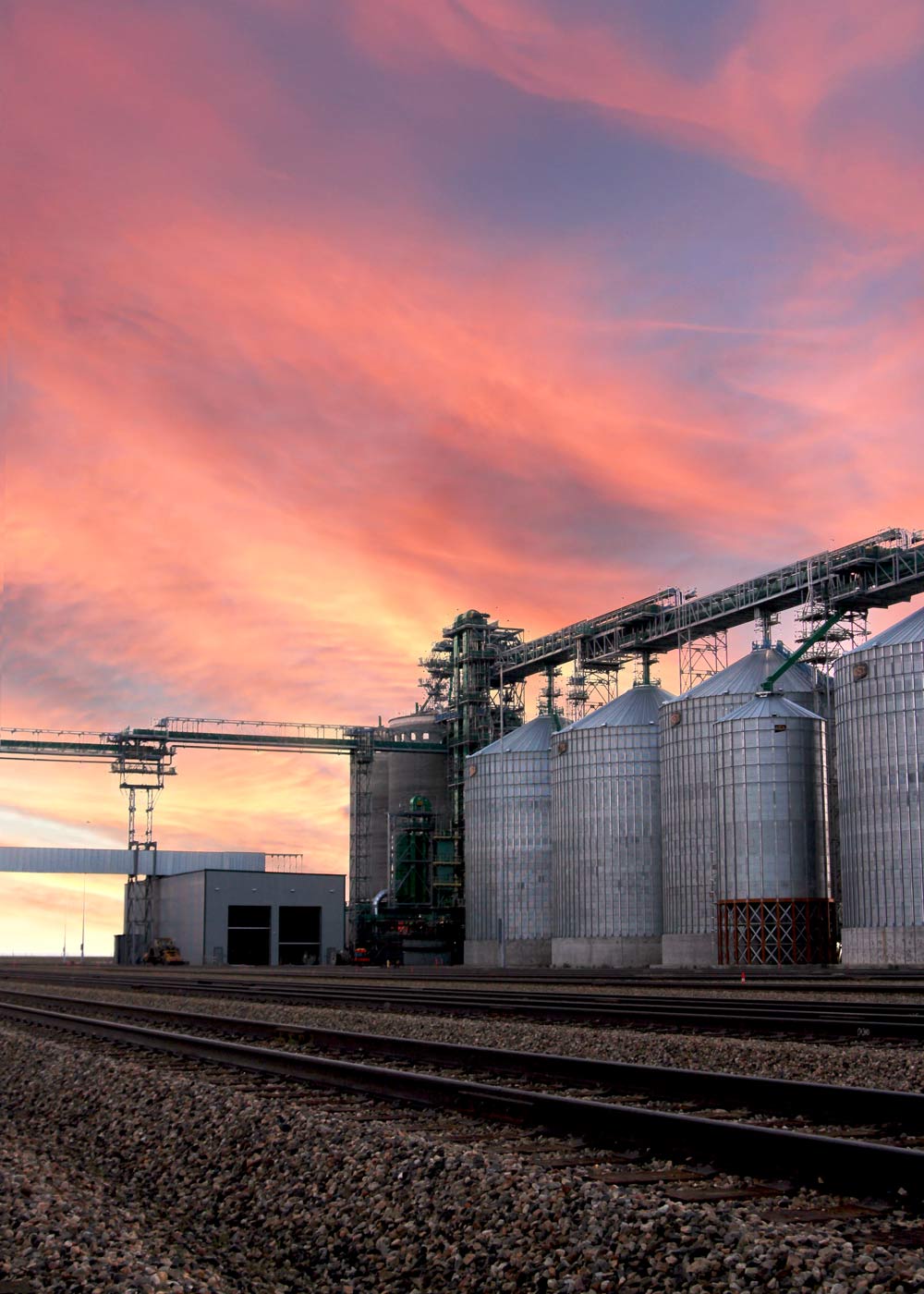 Grain storage silos and rail tracks at an agricultural processing facility at sunset.