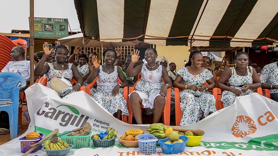 Five women in white dresses sit underneath a striped tent in chairs and wave to the camera while sitting behind a large white Cargill CARE sign and baskets full of fruit. Five women in white dresses sit underneath a striped tent in chairs and wave to the camera while sitting behind a large white Cargill CARE sign and baskets full of fruit.