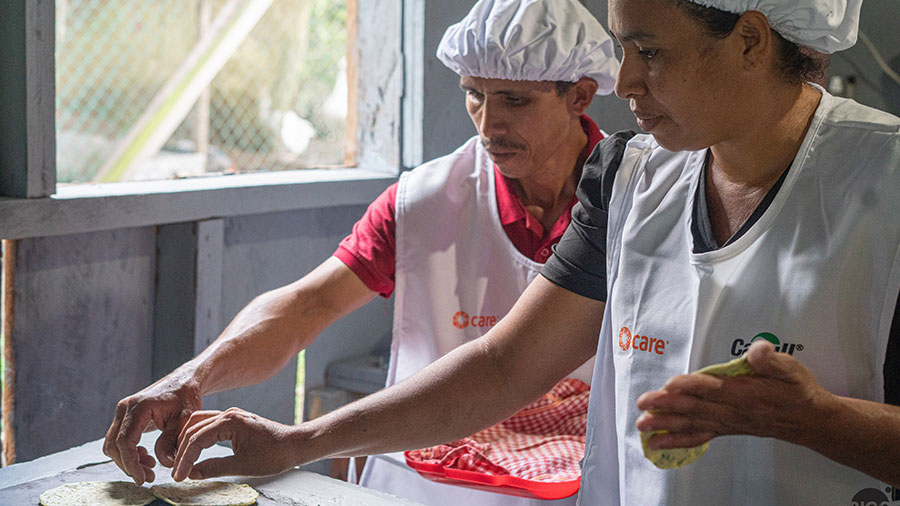 A woman and a man wearing white hair nets and aprons lay down tortillas onto a griddle. 