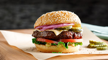 A Cargill TNT burger inside two sesame seed buns with a beef patty, tomatoes, lettuce, pickles, onions and cheese sits on a table. 