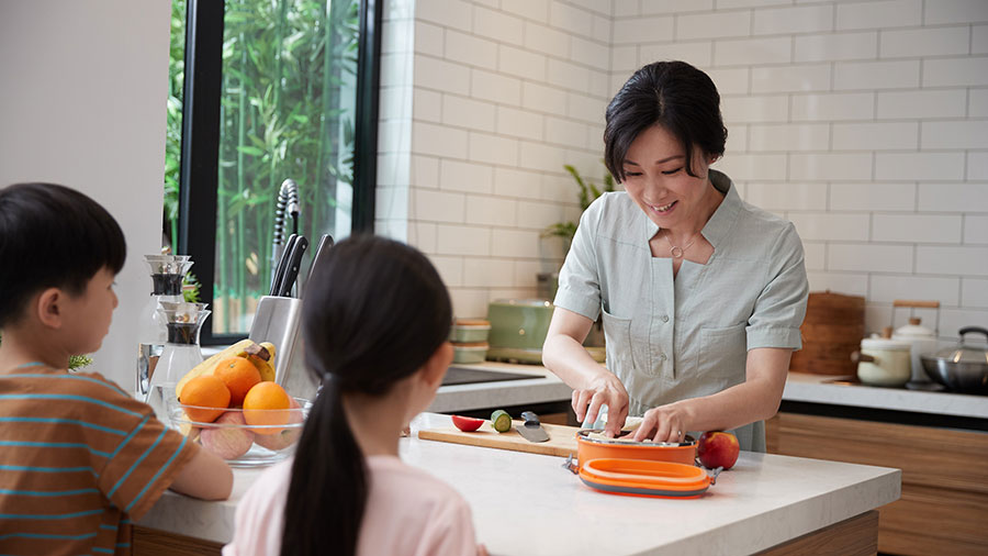 A woman preps a lunch box on a white countertop in a kitchen, while two kids watch from chairs across the countertop. A woman preps a lunch box on a white countertop in a kitchen, while two kids watch from chairs across the countertop.